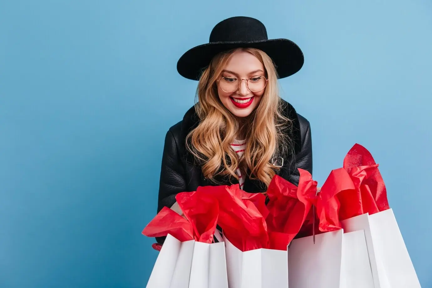 Потрясающая блондинка держит пакеты с покупками. Смеющаяся девушка в элегантной шляпе позирует на фоне синей стены.