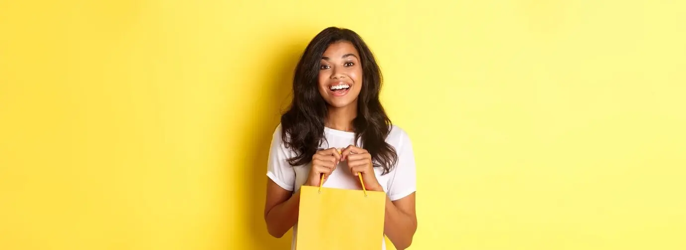 Портрет весёлой афроамериканской девушки, которая во время шопинга открывает пакет с подарком и счастливо улыбается, стоя.